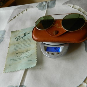 May include: Vintage Ray-Ban sunglasses with green lenses and a gold metal frame. The sunglasses are in a brown leather case and are being weighed on a digital scale. The scale reads 100.0.