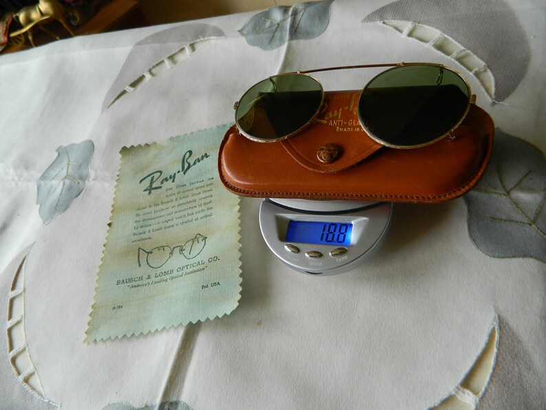 May include: Vintage Ray-Ban sunglasses with green lenses and a gold metal frame. The sunglasses are in a brown leather case and are being weighed on a digital scale. The scale reads 100.0.