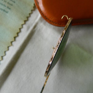 May include: Close-up of a gold-rimmed pair of sunglasses with green lenses. The sunglasses are resting on a white surface.