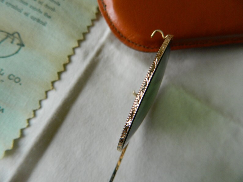 May include: Close-up of a gold-rimmed pair of sunglasses with green lenses. The sunglasses are resting on a white surface.