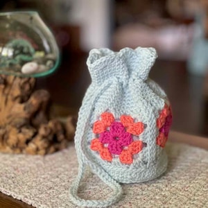 May include: A light blue crocheted drawstring bag with a pink and orange floral design. The bag has a long strap.