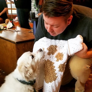 May include: A white and brown crocheted blanket with a paw print design. A person is holding the blanket and looking at a white dog with brown eyes.