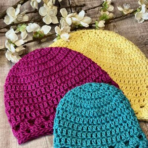 May include: Three handmade crochet beanies in bright yellow, magenta, and teal. Each hat features a consistent textured stitch pattern with a decorative openwork border along the bottom edge. The beanies are arranged in a layered, overlapping stack.