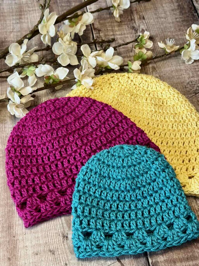 May include: Three handmade crochet beanies in bright yellow, magenta, and teal. Each hat features a consistent textured stitch pattern with a decorative openwork border along the bottom edge. The beanies are arranged in a layered, overlapping stack.