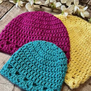 May include: Three handmade crochet beanies in vibrant solid colors: one magenta, one bright yellow, and one teal. Each hat features a consistent, open-stitch pattern crafted from yarn, showcasing a rounded crown shape and a simple, unadorned edge.