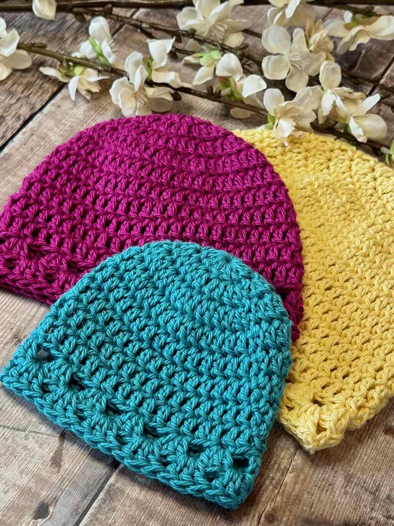 May include: Three handmade crochet beanies in vibrant solid colors: one magenta, one bright yellow, and one teal. Each hat features a consistent, open-stitch pattern crafted from yarn, showcasing a rounded crown shape and a simple, unadorned edge.