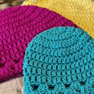 May include: Three crocheted beanies in bright colors, including a teal hat in the foreground, a magenta hat in the middle, and a yellow hat in the back. Each hat features a consistent textured stitch pattern and a rounded crown shape.