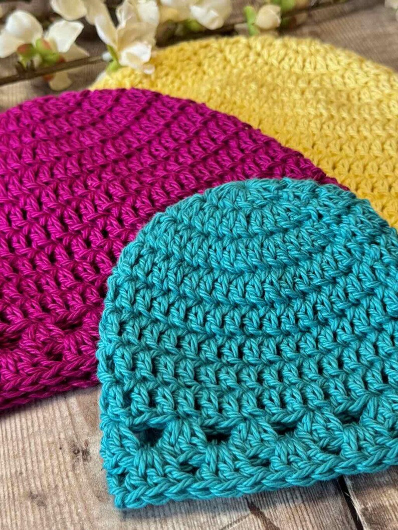 May include: Three crocheted beanies in bright colors, including a teal hat in the foreground, a magenta hat in the middle, and a yellow hat in the back. Each hat features a consistent textured stitch pattern and a rounded crown shape.