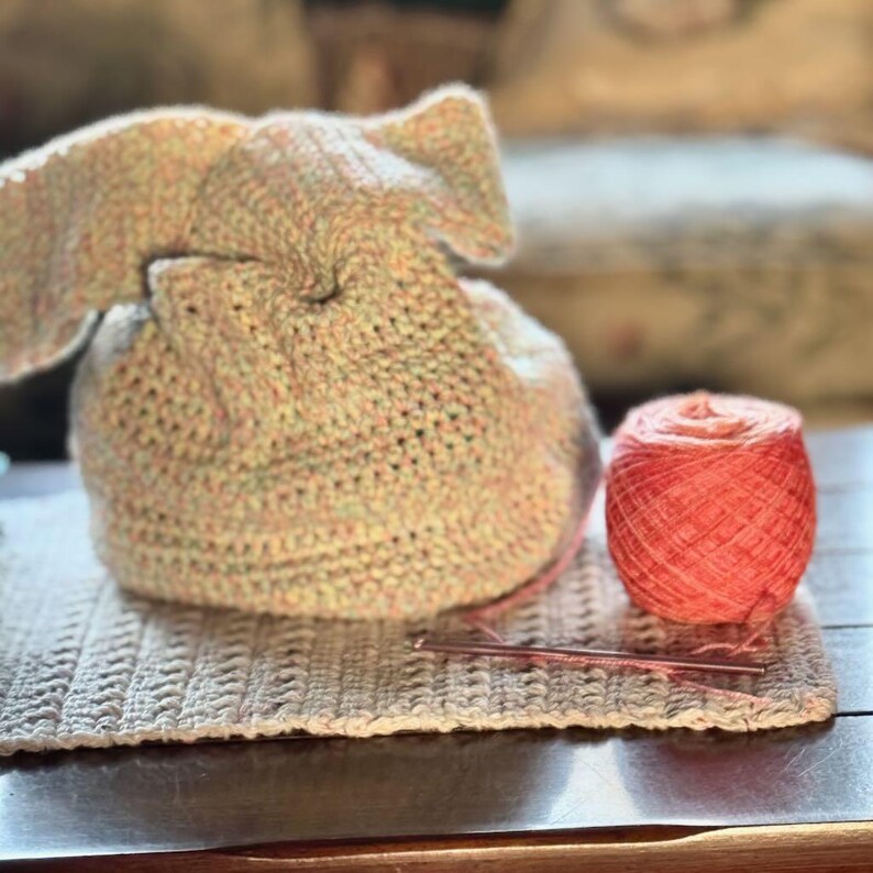May include: A crocheted bunny-shaped bag with a pink yarn ball and a crochet hook. The bag is made with a multi-colored yarn in shades of pink, green, and white.