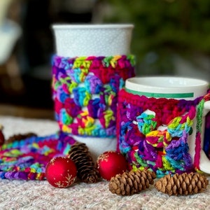 May include: Two mugs with colorful crocheted covers. The covers are made with a variety of colors, including red, pink, purple, blue, green, and yellow. The mugs are sitting on a white surface with some small decorative items.