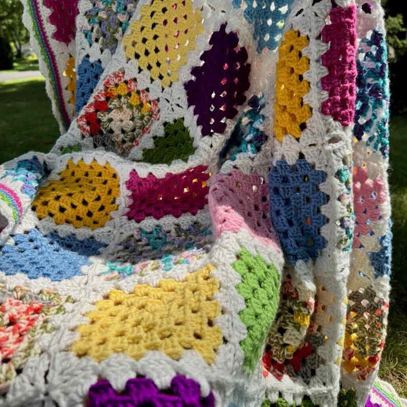 May include: A handmade, colorful crochet blanket. The blanket features a patchwork design with squares in various colors, including yellow, pink, blue, green, and purple, all set against a white background. The blanket is draped over a surface.