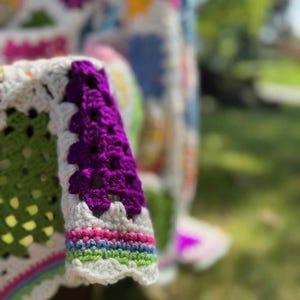 May include: Close-up of a colorful, handmade crochet blanket. The blanket features a white base with green, purple, pink, blue, and yellow squares. The sleeve of the blanket is visible, with a purple cuff and rainbow trim.