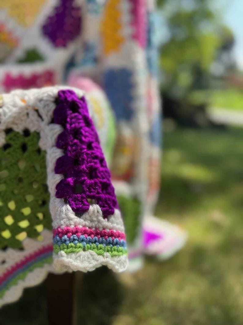 May include: Close-up of a colorful, handmade crochet blanket. The blanket features a white base with green, purple, pink, blue, and yellow squares. The sleeve of the blanket is visible, with a purple cuff and rainbow trim.