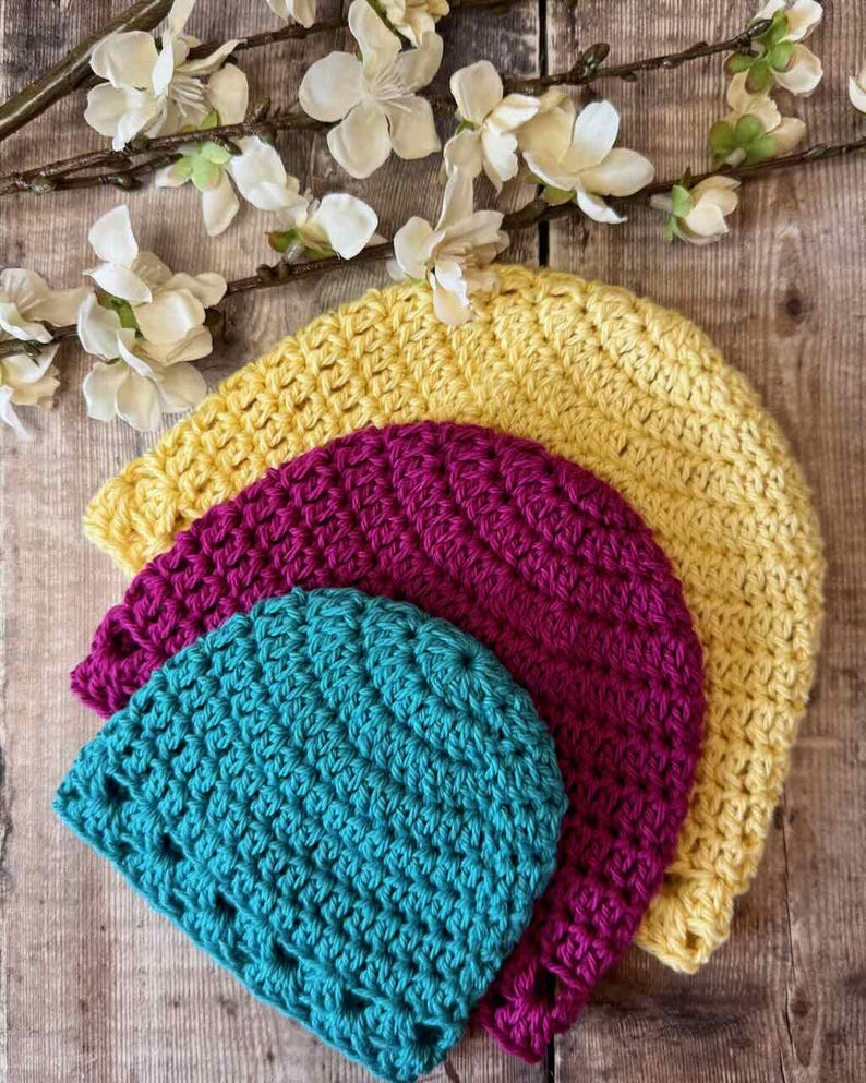 May include: Three crocheted beanies stacked in a nested arrangement. The largest beanie is yellow, the middle beanie is magenta, and the smallest beanie is teal. Each hat features a consistent textured stitch pattern and a rounded crown shape.