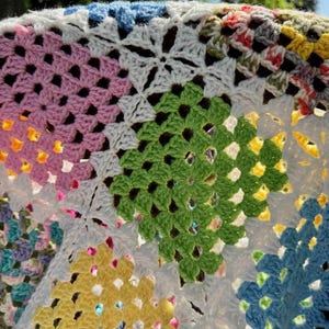 May include: Close-up of a colorful, handmade crochet blanket. The blanket features a patchwork design with various colors, including pink, green, yellow, blue, and white. The crochet stitches create a textured, geometric pattern.