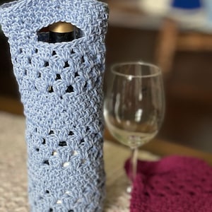 Może przedstawiać: Niebieski, szydełkowy uchwyt na butelkę wina z ozdobnym wzorem. Uchwyt jest wykonany z włóczki i ma otwór na szyjkę butelki.