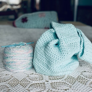 May include: A ball of multi-colored yarn with a crochet hook and a light blue crocheted blanket on a white lace tablecloth.