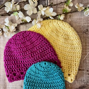 May include: Three crocheted beanies in bright yellow, magenta, and teal. Each hat features a consistent textured stitch pattern with a decorative openwork border along the bottom edge. The beanies are arranged in a layered stack on a wooden surface.