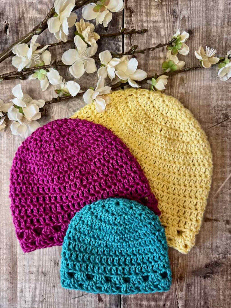 May include: Three crocheted beanies in bright yellow, magenta, and teal. Each hat features a consistent textured stitch pattern with a decorative openwork border along the bottom edge. The beanies are arranged in a layered stack on a wooden surface.