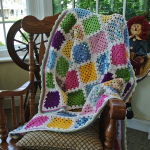 May include: A colorful, handmade crochet blanket draped over a wooden rocking chair. The blanket features a patchwork design with squares of white, green, pink, yellow, blue, and purple. The chair is positioned near a window, with a decorative doll visible.