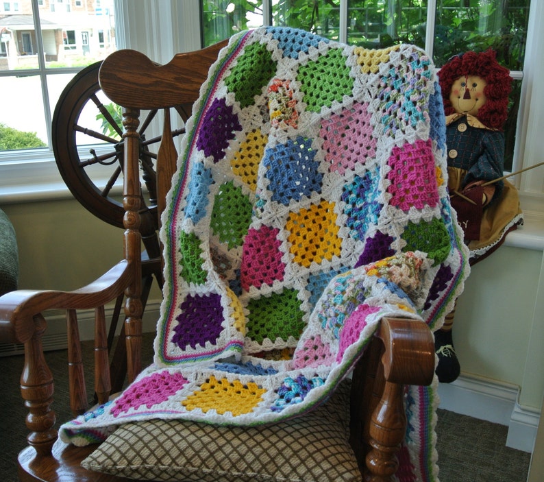 May include: A colorful, handmade crochet blanket draped over a wooden rocking chair. The blanket features a patchwork design with squares of white, green, pink, yellow, blue, and purple. The chair is positioned near a window, with a decorative doll visible.