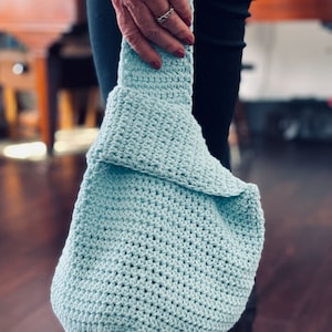 May include: A light blue crocheted tote bag with a slouchy design. The bag is held by a person wearing black pants and boots.