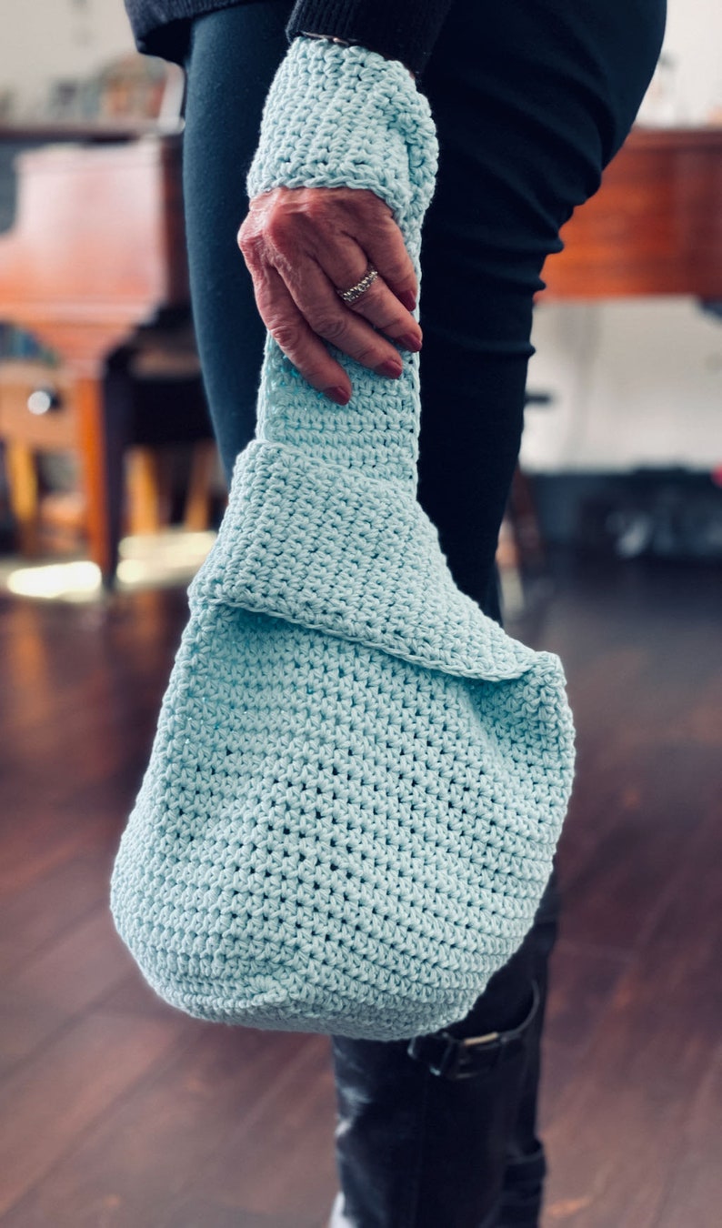 May include: A light blue crocheted tote bag with a slouchy design. The bag is held by a person wearing black pants and boots.
