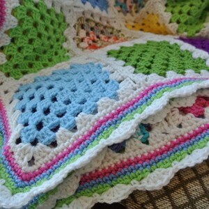 May include: A handmade crochet blanket featuring a patchwork design with squares in various colors, including green, blue, and pink. The blanket has a white scalloped edge with a striped border in pastel colors. The blanket is folded, showing its texture and design.