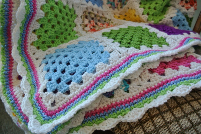 May include: A handmade crochet blanket featuring a patchwork design with squares in various colors, including green, blue, and pink. The blanket has a white scalloped edge with a striped border in pastel colors. The blanket is folded, showing its texture and design.