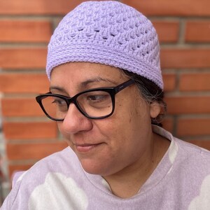 Pu&ograve; includere: Primo piano di una persona che indossa un berretto di lana color lilla chiaro. Il berretto ha un semplice motivo a maglia e viene indossato sulla testa della persona.
