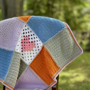 May include: A crocheted baby blanket with a patchwork design in shades of lavender, blue, green, and orange. The center square features a white crocheted heart with a pink center.