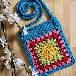 May include: A turquoise blue crochet bag with a long strap. The bag features a central floral design in yellow, pink, red, and green. The bag is handmade and square-shaped. The bag is displayed on a wooden surface with white flowers.