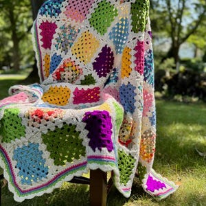 May include: A colorful, handmade crochet blanket draped over a chair. The blanket features a patchwork design with squares in various colors, including pink, green, yellow, and blue, set against a white background. The edges are trimmed with a scalloped border.