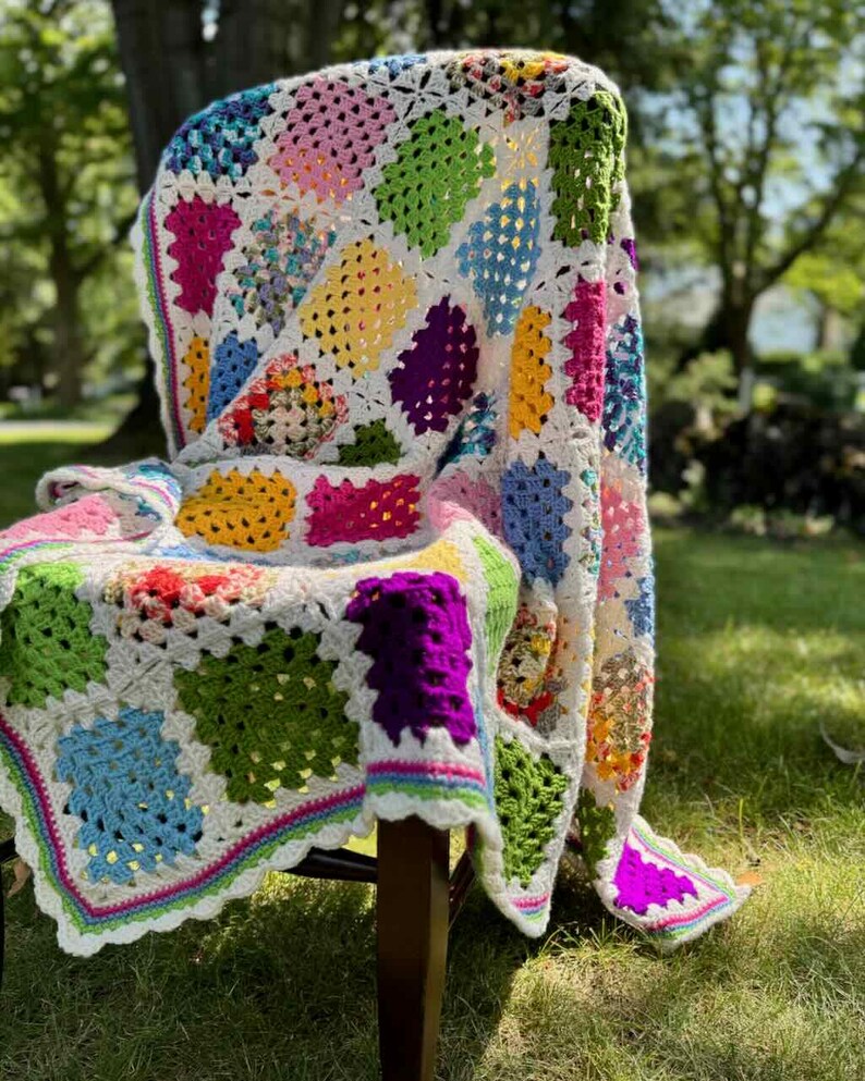 May include: A colorful, handmade crochet blanket draped over a chair. The blanket features a patchwork design with squares in various colors, including pink, green, yellow, and blue, set against a white background. The edges are trimmed with a scalloped border.