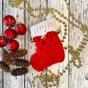 May include: A red and white crocheted Christmas stocking with bells hanging from the top. The stocking is surrounded by gold beads and red ornaments.