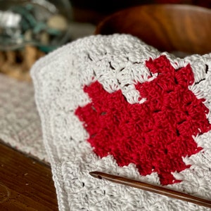 Puede incluir: Una manta de crochet blanca con un diseño de corazón rojo en el centro. Un gancho de crochet de madera está sobre la manta.