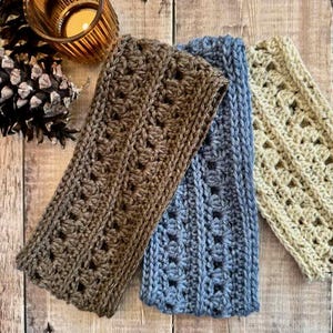 Pode incluir: Três faixas de cabelo de crochê feitas à mão em marrom, azul e bege. As faixas têm padrões texturizados e são exibidas em uma superfície de madeira. Uma vela acesa em um suporte dourado e um pinheiro estão no fundo.