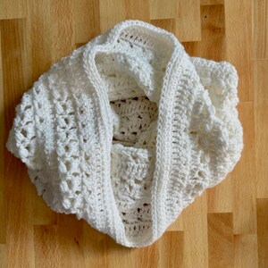May include: White crocheted scarf with a delicate, lacy pattern. The scarf is folded in half and laid flat on a wooden surface.