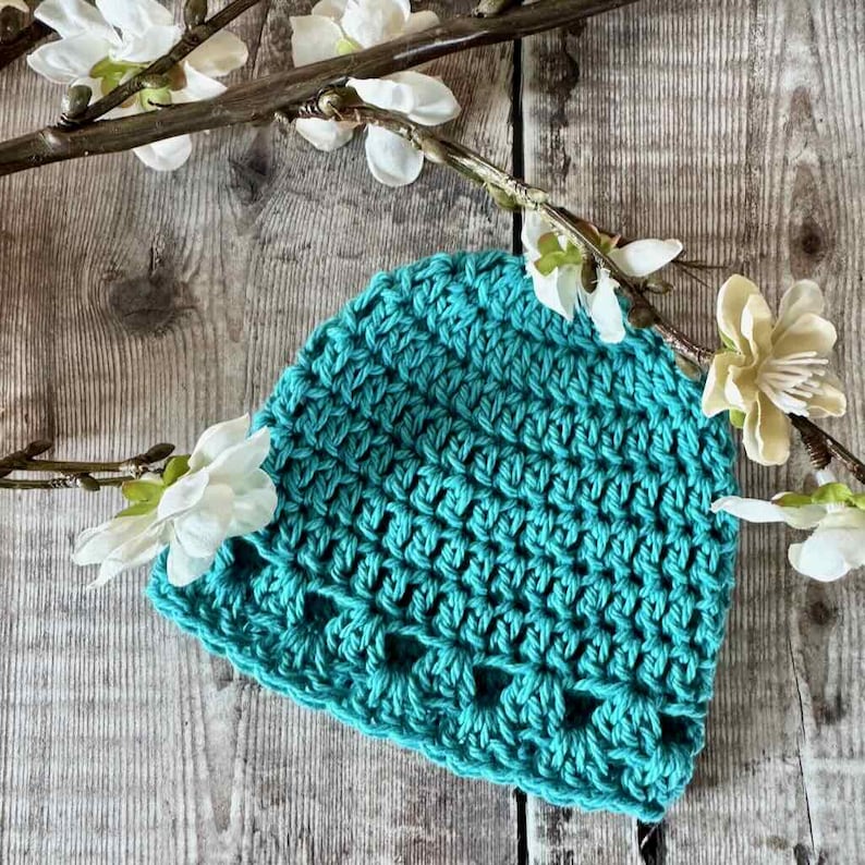 May include: A teal crochet beanie featuring a textured knit pattern with a decorative openwork border along the bottom edge. The hat is displayed with white artificial flowers on a branch, showcasing the vibrant color and detailed yarn construction.
