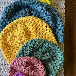 Può includere: Quattro cappelli all'uncinetto in diversi colori: blu, giallo, verde e rosa. I cappelli sono impilati uno sopra l'altro, con il cappello blu in cima e il cappello rosa in fondo.