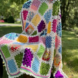 May include: A colorful, handmade crochet blanket draped over a chair. The blanket features a patchwork design with various bright colors, including pink, green, yellow, and blue, set against a white background. The edges are finished with a scalloped border.