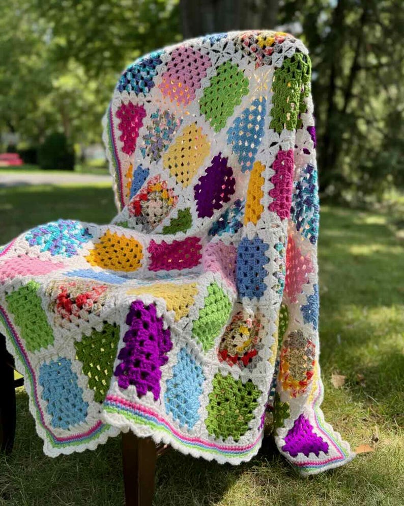 May include: A colorful, handmade crochet blanket draped over a chair. The blanket features a patchwork design with various bright colors, including pink, green, yellow, and blue, set against a white background. The edges are finished with a scalloped border.