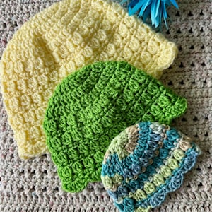 Può includere: Tre cappelli all'uncinetto in diversi colori e motivi. Il cappello più grande è giallo, il cappello intermedio è verde e il cappello più piccolo è blu, verde e beige.