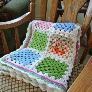May include: A colorful, handmade crochet blanket with a granny square design. The blanket features squares of pink, green, blue, and orange, bordered by white and a scalloped edge. It is draped over a wooden rocking chair.