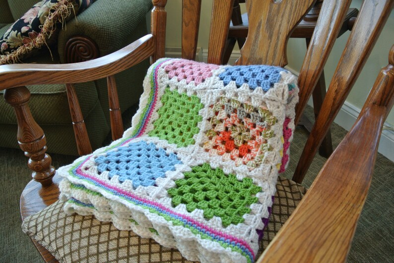 May include: A colorful, handmade crochet blanket with a granny square design. The blanket features squares of pink, green, blue, and orange, bordered by white and a scalloped edge. It is draped over a wooden rocking chair.