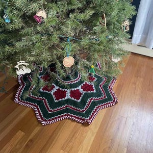 May include: A star-shaped crochet Christmas tree skirt featuring a dark green base with deep red and light gray geometric accents. The edges are finished with a decorative white and red twisted border, placed under a pine tree with ornaments.