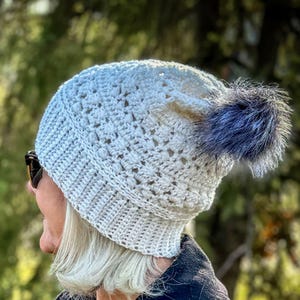 May include: A white crocheted beanie with a blue faux fur pom pom. The beanie has a lacy stitch pattern.