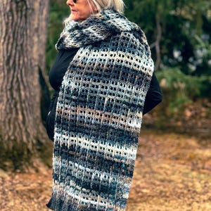 May include: A long, crocheted scarf in shades of gray, brown, and white. The scarf has a lacy, openwork pattern and is worn by a person in a forest setting.