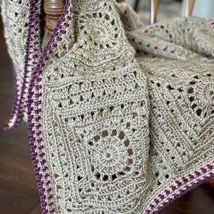 May include: A gray crocheted blanket with a square pattern and a purple border. The blanket is draped over a wooden chair, showcasing its intricate design and texture. The border features a purple and pink trim.