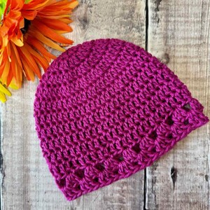 May include: A vibrant magenta beanie crafted with a textured crochet stitch pattern. The hat features a rounded crown and a repeating openwork design along the bottom edge, showcasing a consistent yarn weight and deep, saturated pink color throughout.